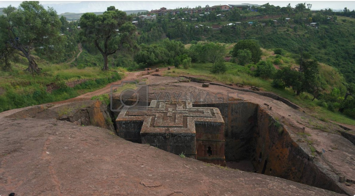 a top view of the church of St George in Lalibela, Ethiopia. a monolithic church carved directly into the earth in hte shape of a cross, surrounded by trenches and lush landscape.